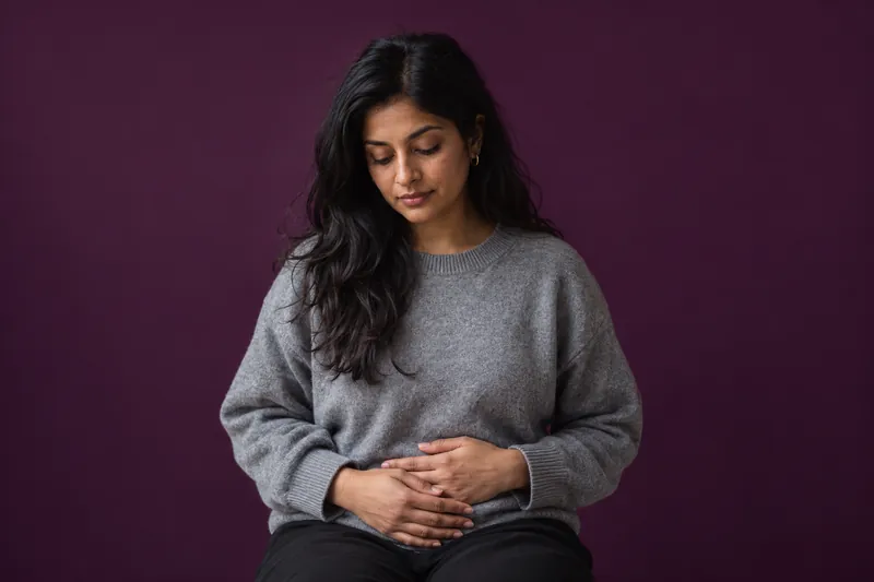 A young woman in a grey knit sweater sits with her hands resting on her stomach and her head bowed, against deep aubergine paper.
