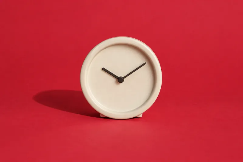 A round cream ceramic table clock with plain hands and no numbers, on cherry-red paper.