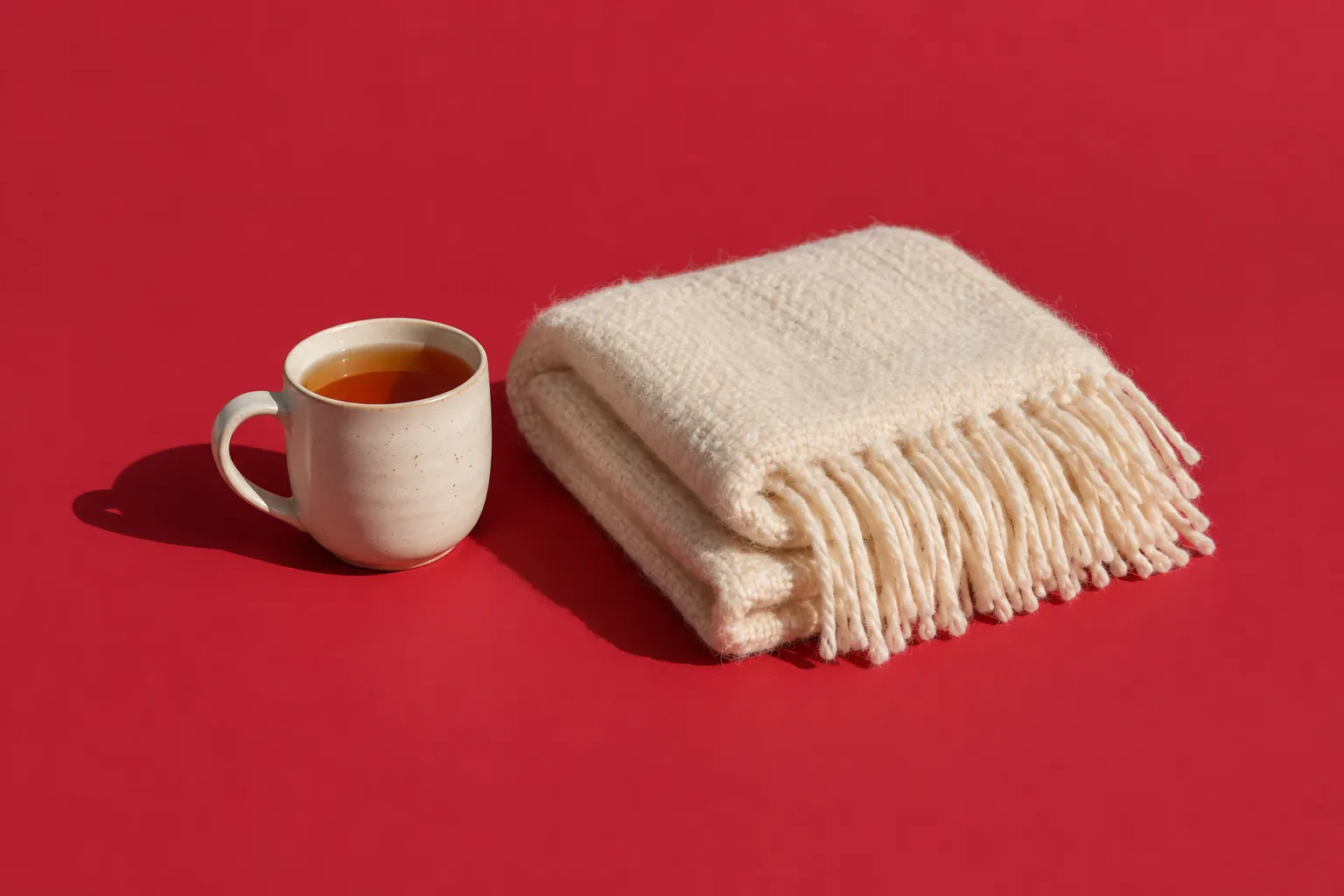 A mug of tea beside a folded cream wool blanket with fringe, on cherry-red paper.