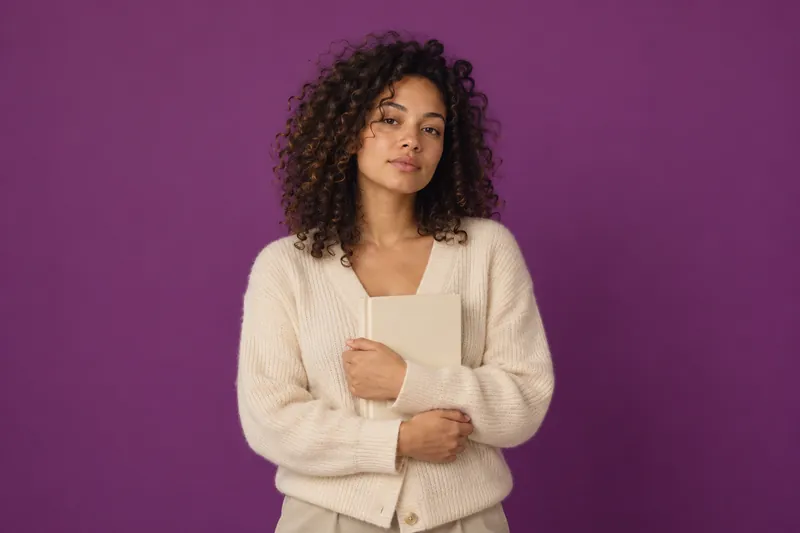 A young woman with curly hair in a cream cardigan holds a plain closed notebook against her chest, against plum paper.