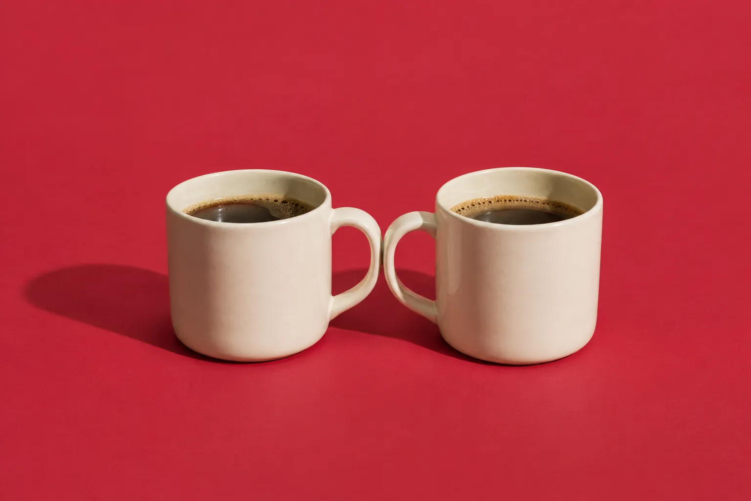 Two matching cream mugs side by side with their handles touching, on cherry-red paper.