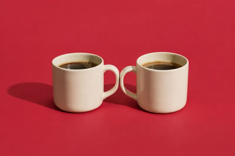 Two matching cream mugs side by side with their handles touching, on cherry-red paper.