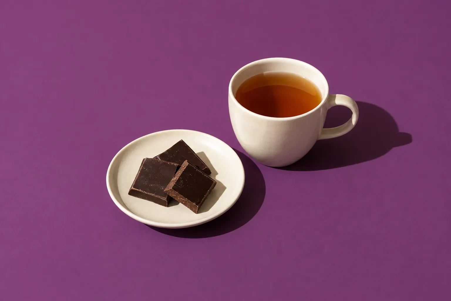 Three squares of dark chocolate on a cream plate beside a cup of tea, on plum paper.