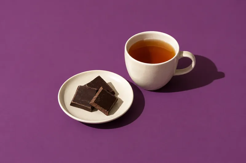 Three squares of dark chocolate on a cream plate beside a cup of tea, on plum paper.