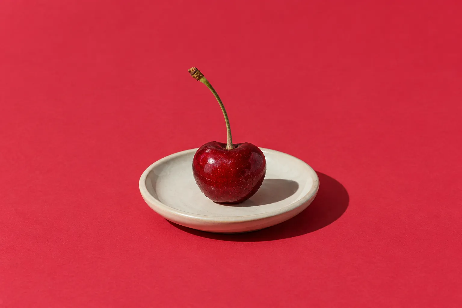 A single ripe cherry with its stem on a small cream saucer, on cherry-red paper.