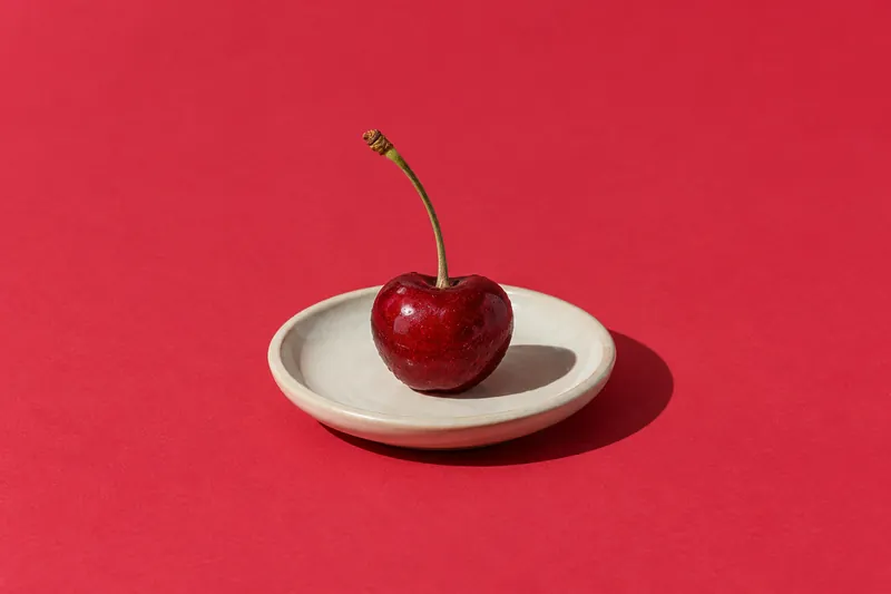 A single ripe cherry with its stem on a small cream saucer, on cherry-red paper.