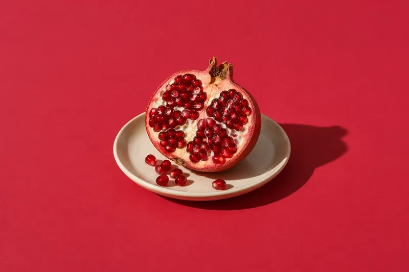 Half a pomegranate on a small cream plate with a few loose seeds, on cherry-red paper.