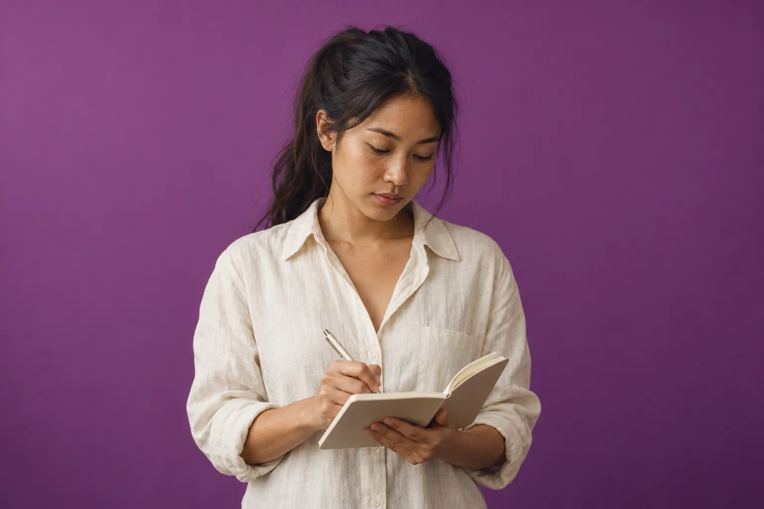 A young woman with a dark ponytail in a linen shirt writes in a notebook, against plum paper.