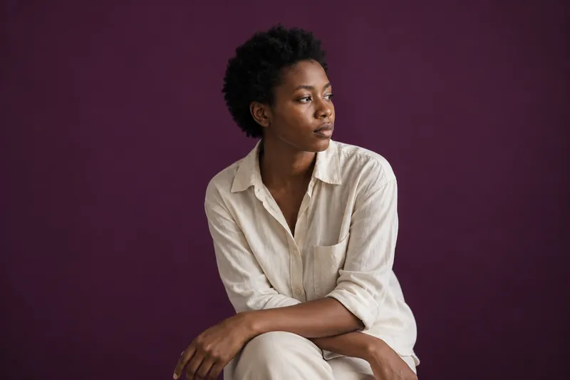 A young woman with short natural hair in a cream shirt sits side-on, looking away, against deep aubergine paper.