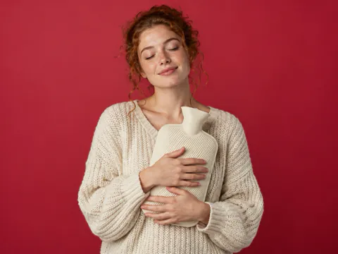 A woman with red hair hugs a knitted hot water bottle against cherry paper, eyes closed.