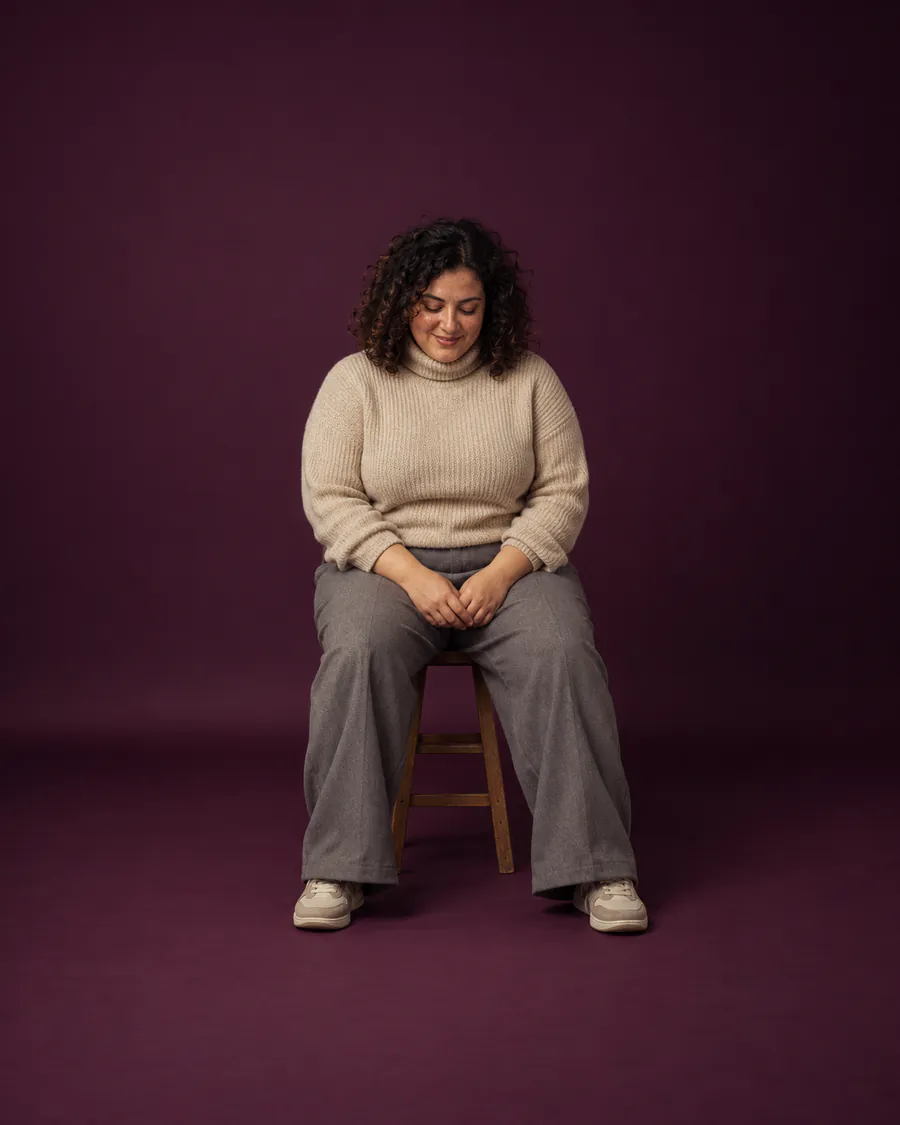 A woman with curly hair and freckles sits on a stool in a pool of warm light against dark aubergine paper, looking down at her hands.