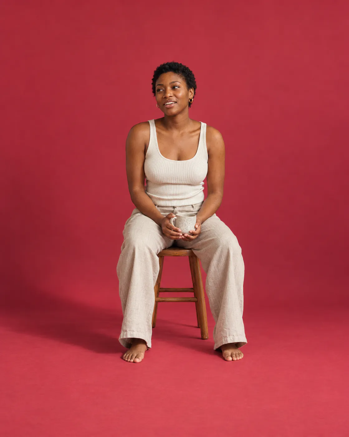 A young woman with short natural hair sits on a stool against cherry-red paper, holding a mug and talking.