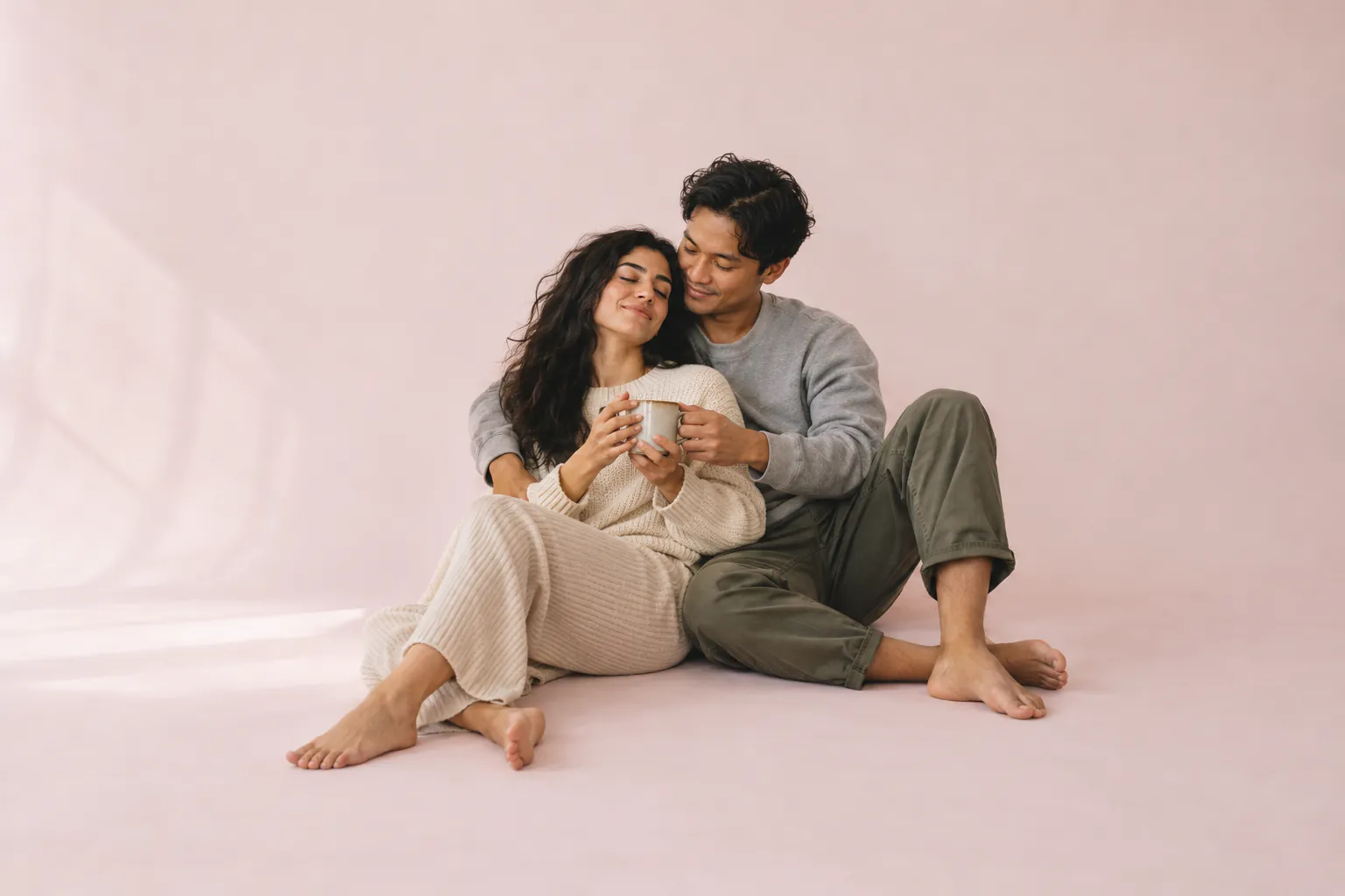 A couple sit together on the floor against pale pink paper. She leans back into him, eyes closed and smiling, while he hands her a mug.