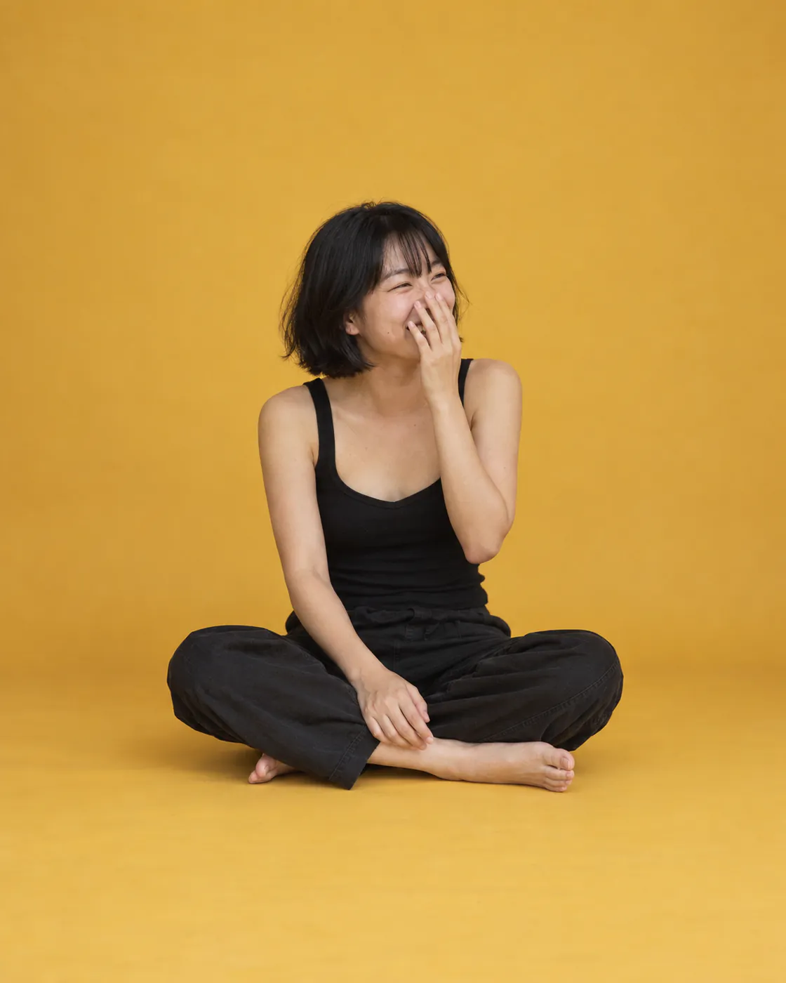 A woman with a black bob sits cross-legged on mustard-yellow paper, laughing with her hand over her mouth.